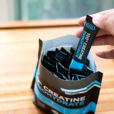 Hand holding a creatine monohydrate supplement stick pack over a package on a wooden surface.