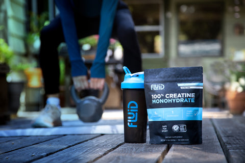 Bag of 100% Creatine Monohydrate and branded shaker bottle from Fluid Sports Nutrition. Woman lifts kettlebell in background.