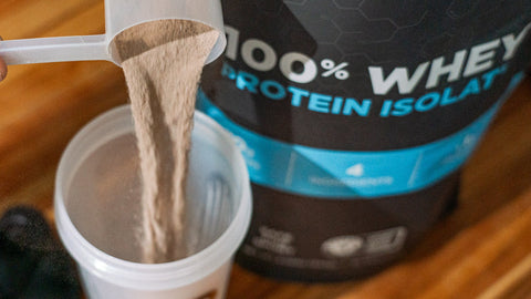 Fluid Sports Nutrition whey protein isolate powder being poured into a shaker bottle, with product in the backdrop.