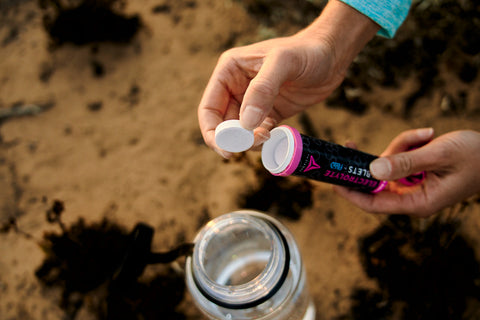 Person holding a water bottle cap and a black and pink container on a sandy background