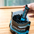 Hand holding a creatine monohydrate supplement stick pack over a package on a wooden surface.