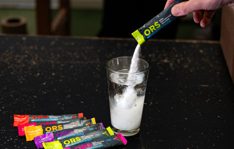 Person dissolving an ORS powder packet into a glass of water on a dark surface.