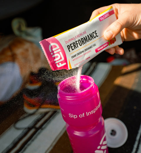 Person pouring Fluid Performance Raspberry Lemonade 50g Carb packet into a pink sports bottle.