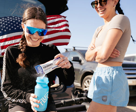 Two women outdoors with an American flag, one holding a Fluid performance drink mix packet and a blue Fluid water bottle.