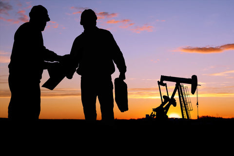 Two silhouetted figures standing next to an oil pump against a sunset sky.